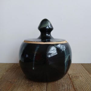 Peut inclure: Un pot en céramique noir avec un couvercle. Le pot a un corps rond et un bouton pointu sur le couvercle. Le pot a une finition brillante avec des stries bleues et vertes. Le pot est posé sur une surface en bois.
