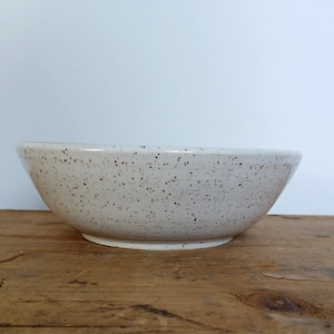 Peut inclure: Un bol en céramique blanc moucheté, de forme large et peu profonde. Le bol a une forme arrondie et une base légèrement surélevée. La surface est couverte de petites mouchetures brun foncé, lui donnant un aspect rustique. Le bol repose sur une surface en bois.