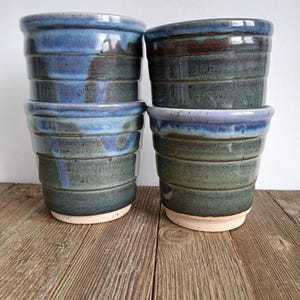 May include: Four ceramic pots in shades of blue and green are stacked on a wooden surface. The pots have a textured, horizontal-lined design and a light-coloured base. The glaze creates a glossy finish, enhancing the colour variations.