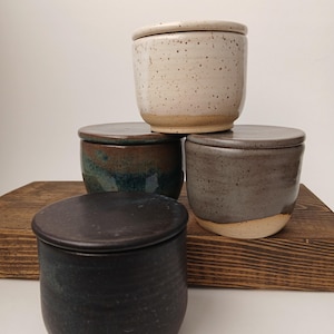 May include: Four ceramic jars with lids arranged in a tiered formation. The top jar is cream with brown speckles. The middle jars are a dark blue-green and a solid grey. The front jar is matte black. Each jar has a cylindrical shape and a flat lid.