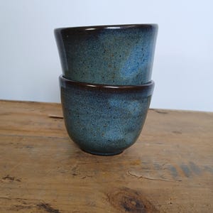 May include: Two handmade ceramic cups stacked on top of each other. The cups have a rounded shape with a speckled blue glaze and a dark brown rim. The cups are on a wooden surface.