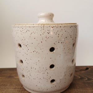 May include: A cylindrical ceramic garlic keeper with a speckled off-white glaze and a matching lid. The body features multiple circular ventilation holes arranged in a grid pattern to allow for airflow, with a small knob handle on the lid.