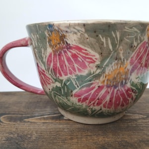 May include: A ceramic mug featuring a hand-painted floral pattern with pink coneflower-like blossoms, green foliage, and orange centers against a speckled grey background. The mug has a curved pink handle and a wide, rounded bowl shape.