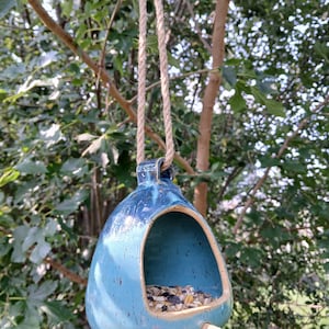 May include: A teardrop-shaped ceramic bird feeder with a glossy blue glaze and dark speckles. It features a large front opening filled with birdseed, a small wooden perch extending from the bottom, and a thick jute rope for hanging.