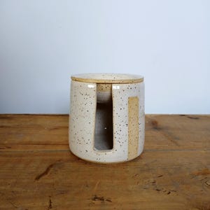May include: A cylindrical ceramic yarn bowl with a speckled cream glaze and a smooth, unglazed vertical rectangular accent on the side. The bowl features a circular lid and a deep, U-shaped cutout on the front rim for guiding yarn through the vessel.