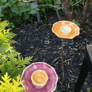 Peut inclure: Deux tuteurs de jardin en céramique avec des sommets en forme de fleur. Une fleur a des pétales orange brillants avec un centre jaune pâle, tandis que l'autre a des pétales magenta brillants avec un centre orange clair, sur des tiges métalliques.