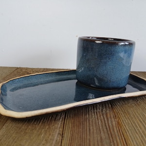 May include: A blue ceramic cup and saucer. The cup is cylindrical with a dark blue glaze and a brown rim. The oblong saucer has a matching blue glaze and a cream edge. The set is displayed on a wooden surface. The cup is approximately 7 cm tall.