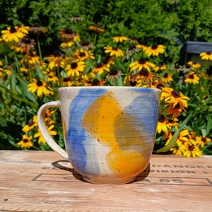 May include: A ceramic mug with a speckled off-white base featuring abstract brushstrokes of bright yellow and blue. The mug has a rounded body, a curved handle on the left side, and a visible horizontal texture from the pottery wheel process.