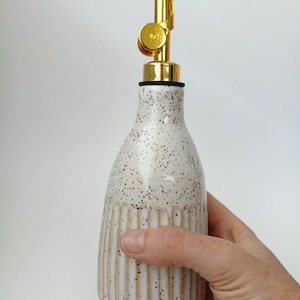 Peut inclure: Un distributeur d'huile en céramique avec un vernis blanc moucheté et une texture nervurée verticale sur la moitié inférieure. La bouteille est dotée d'un bec verseur en métal doré brillant avec un mécanisme de bouchon à charnière sur le dessus.
