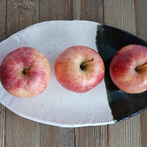 May include: Three red and yellow apples on a white and black ceramic serving dish. The apples are round with a stem and are arranged in a row. The dish has a speckled pattern on the white side.