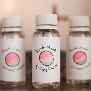 May include: Three clear glass bottles with white lids and labels. The labels have a pink, orange, and blue watercolour design of a moon with the text "Blood Moon Eclipse Water".