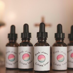 May include: Five small brown glass bottles with dropper tops. Each bottle has a white label with the text "Blood Moon Eclipse Water" and a hand-drawn illustration of a pink and blue moon.