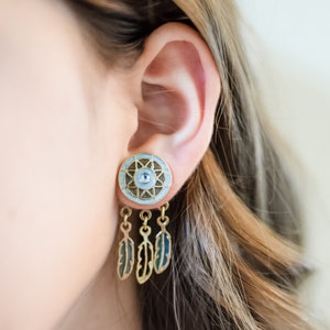 May include: Gold tone earrings with a blue and white circular charm featuring a starburst design and a blue eye in the center. The charm is attached to three gold tone feathers.