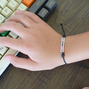 May include: A black string bracelet with a silver charm that says "code like a girl" in pink letters.