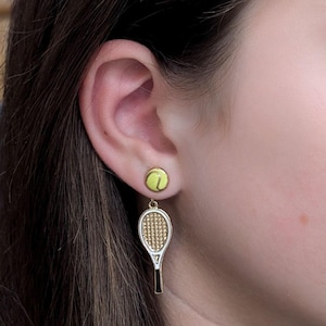 Puede incluir: Un par de pendientes de tenis de oro. Un pendiente presenta una raqueta de tenis con un diseño dorado y negro. El otro pendiente presenta una pelota de tenis amarilla.