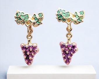 Könnte beinhalten: Goldfarbene Ohrringe mit einem lila Trauben-Design und grünen Blättern. Die Ohrringe sind im Hänge-Stil und haben einen Clipverschluss.