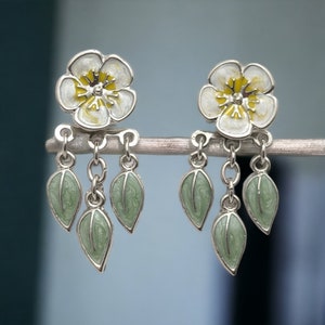 Eye-catching ear jackets showcasing a front design of cherry blossoms in a lovely palette of white and yellow, and a playful back design featuring dangling green leaves. Crafted hypoallergenic and nickel-free, with a hand-painted finish.