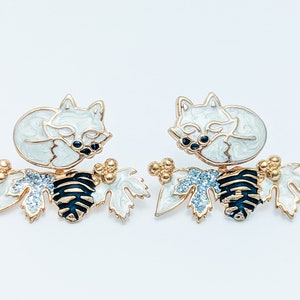 Close-up view of the winter version of the white fox front back earrings, featuring a sleeping white fox and branch with glittery white leaves."