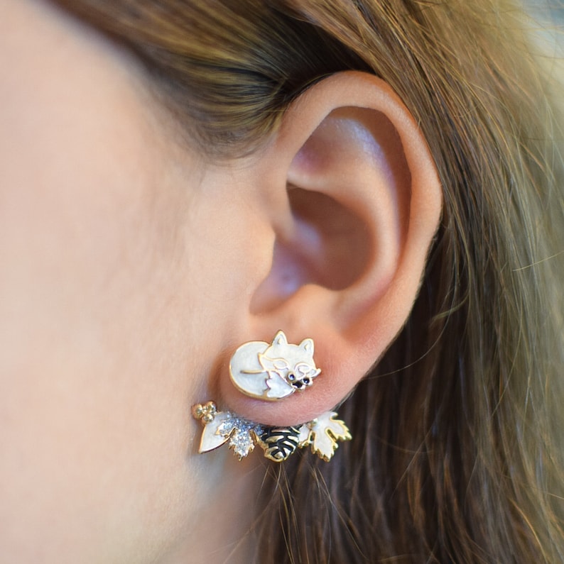 A close-up of a person wearing whimsical front-back earrings, featuring a white fox stud in the front and a golden, glitter-embellished winter leaves and pinecone that peeks out from behind the earlobe.