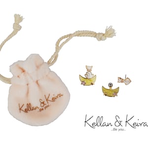 Puede incluir: Un par de pendientes de tono dorado con un dise&ntilde;o de gato blanco sobre una luna creciente. Los pendientes est&aacute;n empaquetados en una peque&ntilde;a bolsa de tela blanca con cord&oacute;n, en la que se imprime el texto "Kellan & Keira...Be you."