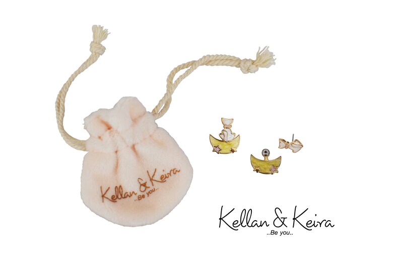 Puede incluir: Un par de pendientes de tono dorado con un dise&ntilde;o de gato blanco sobre una luna creciente. Los pendientes est&aacute;n empaquetados en una peque&ntilde;a bolsa de tela blanca con cord&oacute;n, en la que se imprime el texto "Kellan & Keira...Be you."