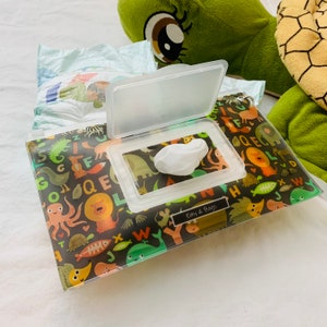 May include: A clear plastic wipe dispenser with a black and white animal print design. The dispenser has a flap that opens to reveal a white wipe. The text "Emy & Bags" is printed on the dispenser.