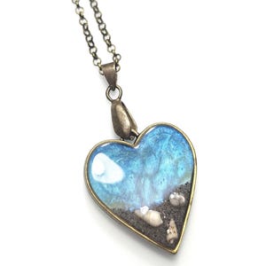 Op de afbeelding: Hartvormige hangerketting met een bronskleurige ketting. De hanger heeft een blauw-witte oceaanscène met zand en schelpen. De hanger is ongeveer 3,8 cm breed.