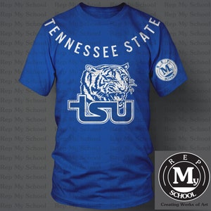 May include: A blue t-shirt with a white tiger graphic and the words "Tennessee State" and "TSU" on the front. The shirt also has a white circle with the letters "REP" and "MY" and the word "SCHOOL" on the right sleeve. The shirt is on a gray background.