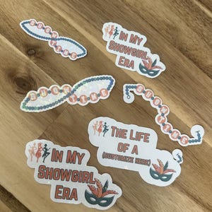 May include: A selection of stickers with a showgirl theme. The stickers display phrases such as "SHOWTIME" and "IN MY SHOWGIRL ERA". Some stickers include a mask with feathers. The stickers are white with a holographic finish.