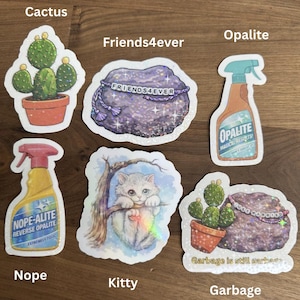 May include: A selection of stickers with diverse designs. One depicts a cactus in a terracotta pot, another says "Friends4ever" on a purple rock. Other stickers include a spray bottle labeled "Opalite" and a kitten.