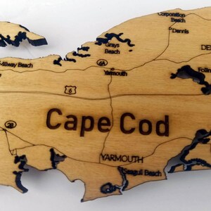 May include: A wooden map of Cape Cod with black outlines of the coastline and towns. The map includes the names of towns and beaches, including Millway Beach, Barnstable, Grays Beach, Corporation Beach, Dennis, Follins Pond, Yarmouth, and Seagull Beach.