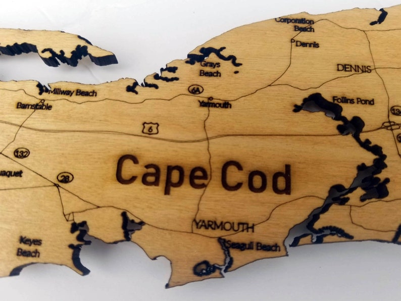 May include: A wooden map of Cape Cod with black outlines of the coastline and towns. The map includes the names of towns and beaches, including Millway Beach, Barnstable, Grays Beach, Corporation Beach, Dennis, Follins Pond, Yarmouth, and Seagull Beach.