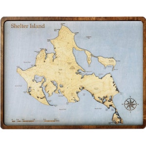 May include: Wooden map of Shelter Island with a light blue background and a dark brown frame. The map shows the island's topography and geographical features, with the text "Shelter Island" at the top.