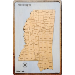 May include: A wooden map of Mississippi with the state's counties outlined. Each county is labeled with its name. The map is on a light blue background with a wooden frame.