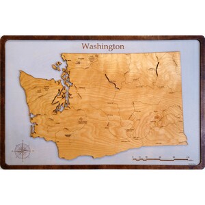 May include: A wooden map of the state of Washington, with the state name printed at the top. The map is mounted on a light gray background with a dark brown frame. A compass rose is in the lower left corner. The map includes a scale in miles.
