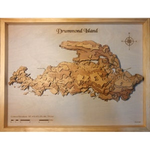 May include: A wooden relief map of Drummond Island, showing contour lines and the island's topography. The map is framed in a light wood frame and includes a compass rose and a scale bar.