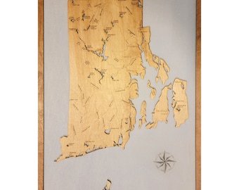 Op de afbeelding: Een houten kaart van Rhode Island met de naam van de staat bovenaan en een kompasroos rechtsonder. De kaart is gemonteerd op een lichtbruin houten frame. De kaart toont de omtrek van de staat met de namen van steden en dorpen binnen de staat.