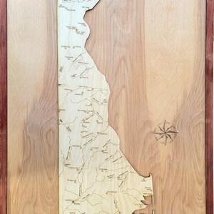 May include: Wooden map of Delaware, laser-cut with detailed city and river outlines. The map is set within a dark wood frame, with the state name carved at the top. A compass rose is etched on the right side.