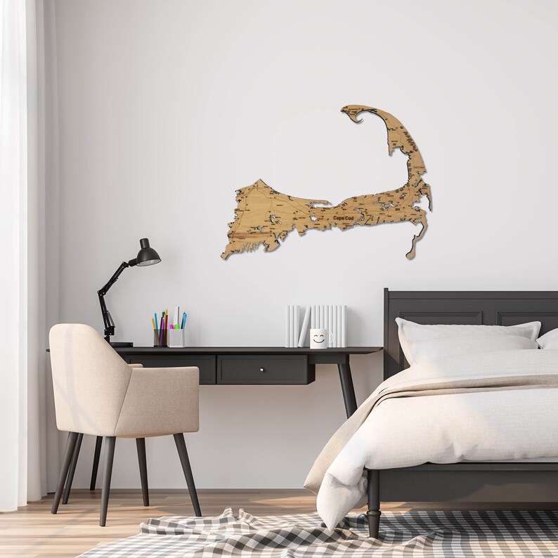May include: A wooden map of Cape Cod, Massachusetts, mounted on a white wall in a bedroom setting. The map is a light brown color and has a detailed outline of the Cape Cod peninsula. The map is hanging above a black desk with a lamp and a chair. There is a bed with white bedding in the background.