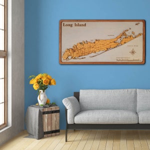 May include: A wooden map of Long Island, New York, with a light brown frame. The map is mounted on a light blue wall above a light gray couch. A vase of yellow roses sits on a wooden table in front of the couch.