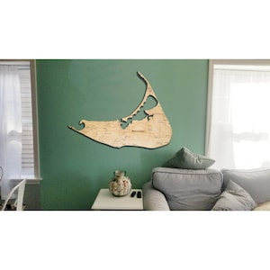 May include: A wooden map of Nantucket Island, a popular vacation destination, is mounted on a green wall. The map is a light brown color and has a detailed outline of the island. The word "Nantucket" is printed in the center of the map.