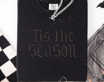 May include: Black sweatshirt with the phrase "Tis the Season" in gothic lettering, with bat graphics. A silver chain necklace is draped across the top. The sweatshirt is folded, with a Comfort Colors tag visible.