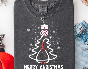 May include: Dark gray t-shirt with a white Christmas tree design incorporating a nurse's hat, stethoscope, and heart. The words "MERRY CHRISTMAS" are printed in white at the bottom. A silver necklace with a sun pendant is visible.