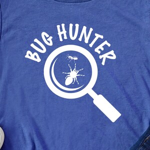Puede incluir: Una camiseta azul con el texto "BUG HUNTER" en blanco, que presenta una lupa con una araña y una hormiga dentro.