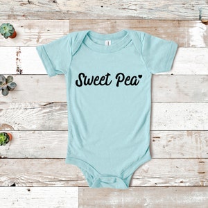 Może przedstawiać: Jasnoniebieskie body niemowlęce z napisem "Sweet Pea" w czarnym skrypcie, z sercem. Ubranko jest umieszczone na jasnym drewnianym tle, z małymi sukulentami w doniczkach.