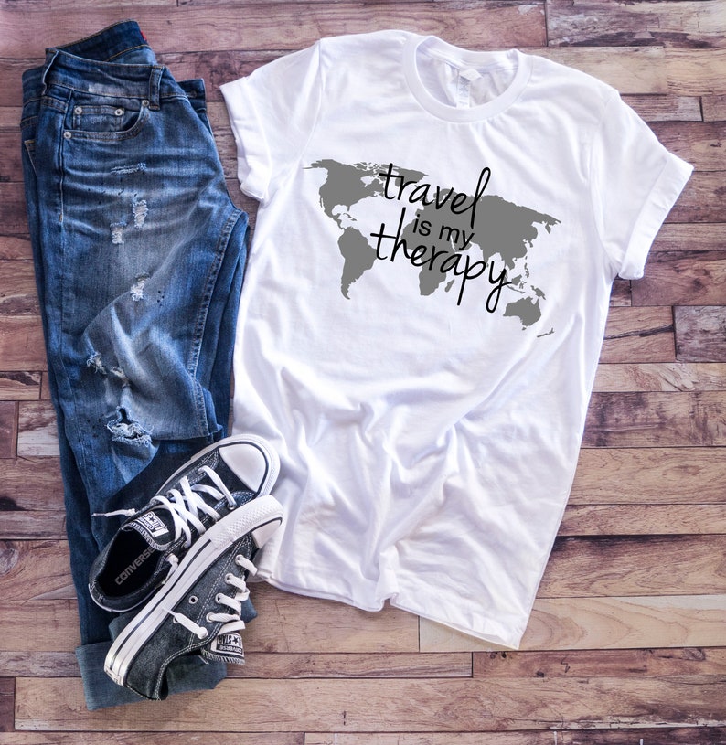 May include: White t-shirt with a gray world map graphic and the text "travel is my therapy" in black.