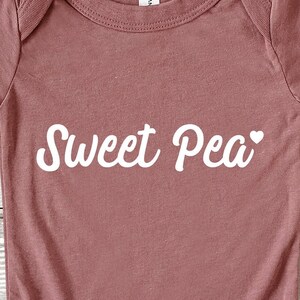 Może przedstawiać: R&oacute;żowy kombinezon dla dziecka z napisem "Sweet Pea" w białym skrypcie. Małe białe serce znajduje się obok słowa "Pea". Kombinezon jest wykonany z miękkiego materiału.