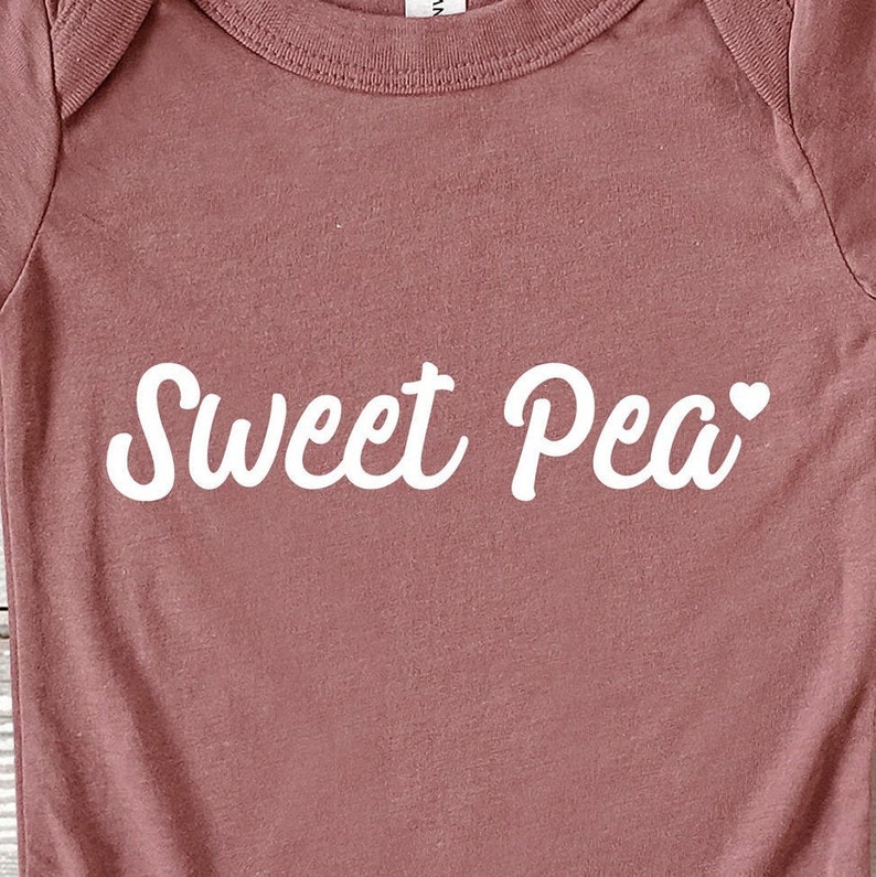 Może przedstawiać: R&oacute;żowy kombinezon dla dziecka z napisem "Sweet Pea" w białym skrypcie. Małe białe serce znajduje się obok słowa "Pea". Kombinezon jest wykonany z miękkiego materiału.