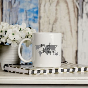 May include: White ceramic mug with a gray outline of the world and the text "travel is my therapy".
