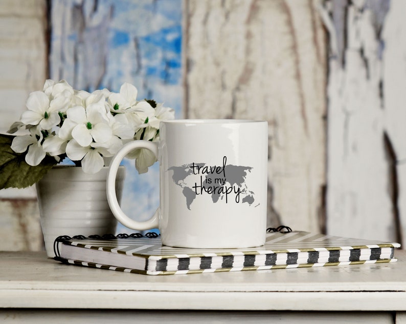May include: White ceramic mug with a gray outline of the world and the text "travel is my therapy".